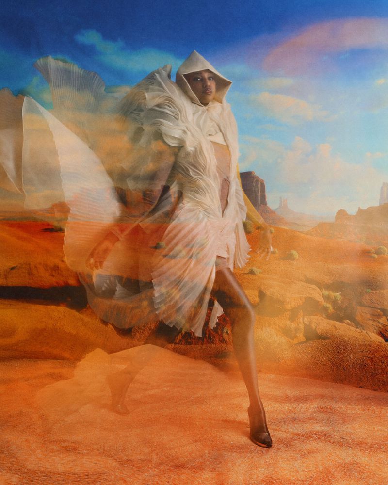 Model wearing a sculpted, off-white hooded pleated garment layered over an arid orange desert landscape with a blue sky.