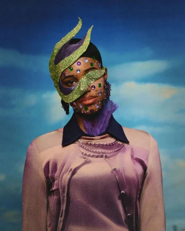 Model wearing an ornate green and purple rhinestone mask, crystal face embellishments, and a lilac top before a blue sky backdrop.