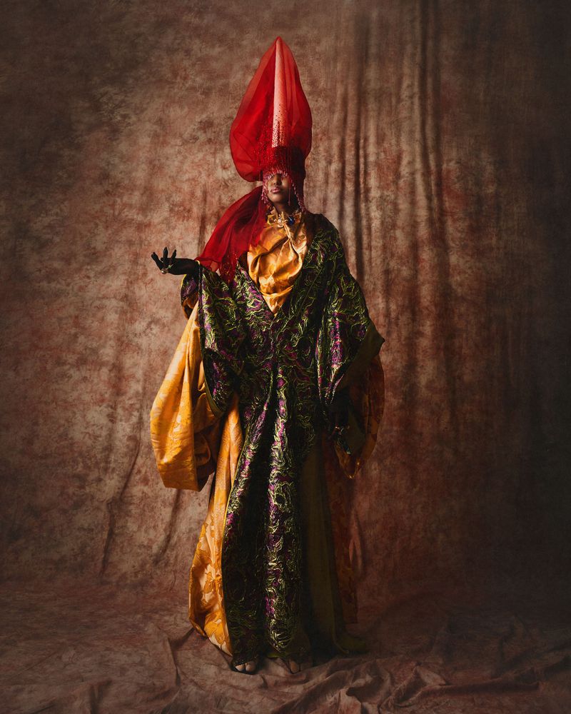 Model in an elaborate green brocade robe and tall red headdress posing against a mottled brown textured backdrop.