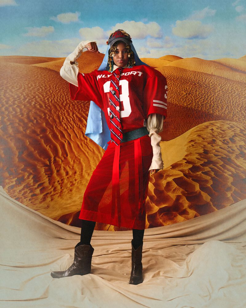 Model flexing in a red numbered football jersey, sheer red skirt, striped tie, and boots in front of a desert backdrop.