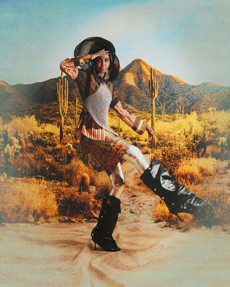 Model in an eclectic patterned ensemble and slouchy black boots posing dynamically against a desert landscape backdrop.