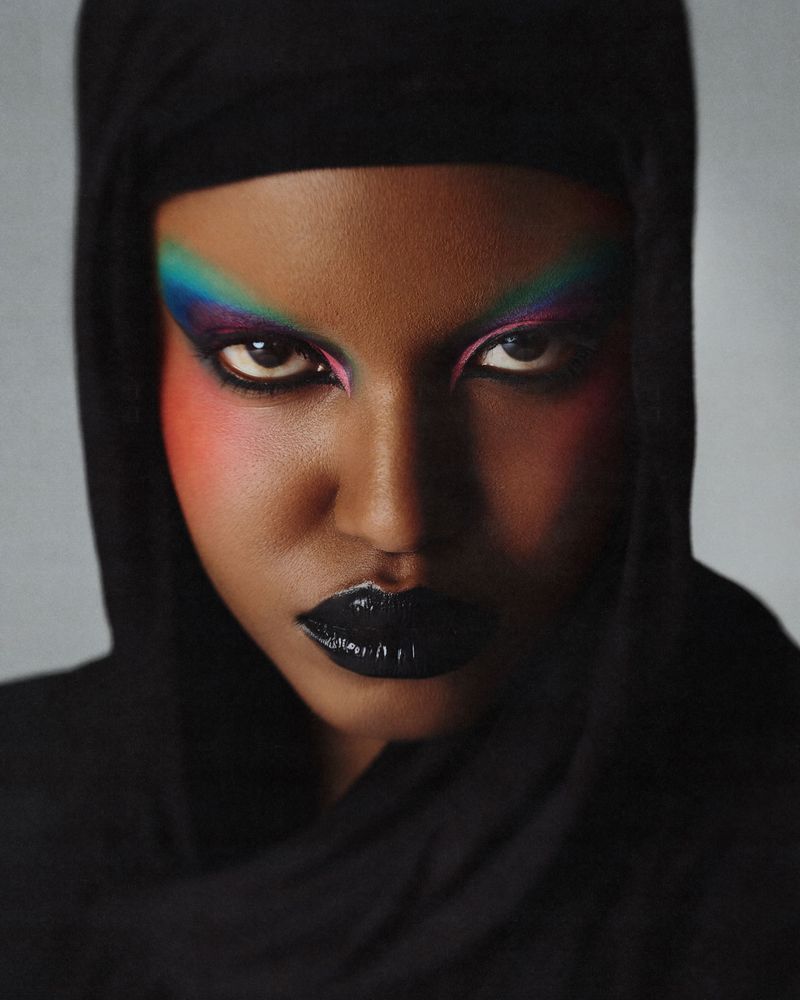 Close-up portrait of a model with dramatic colorful eye makeup, glossy black lipstick, and a black hood framing the face.