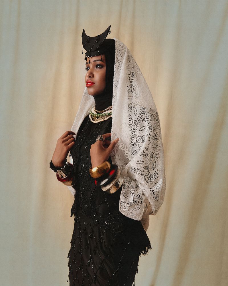 Model wearing a black beaded ensemble, white lace veil, and crescent headpiece posing against a neutral draped backdrop.