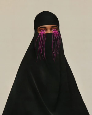 Portrait of a person wearing a black niqab with dramatic long magenta fringe eyelashes against a neutral studio background.