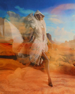 Model wearing a sculpted, off-white hooded pleated garment layered over an arid orange desert landscape with a blue sky.