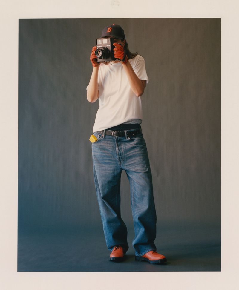 Subject holding a vintage camera in front of their face, wearing a white t-shirt, baggy denim jeans, cap, and gloves in a studio.