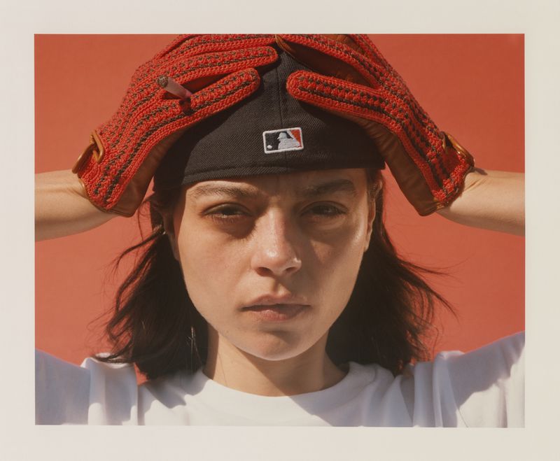 Portrait of a woman wearing red crocheted gloves on her head holding a cigarette, with a red background and white border.