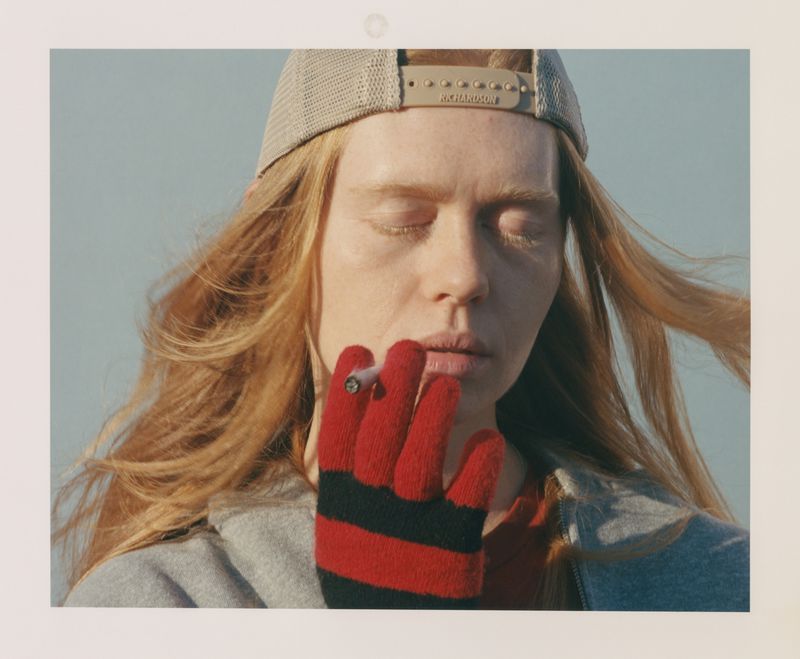 Close-up of a person wearing a backward trucker cap and striped glove holding a smoking joint with closed eyes against an open sky.