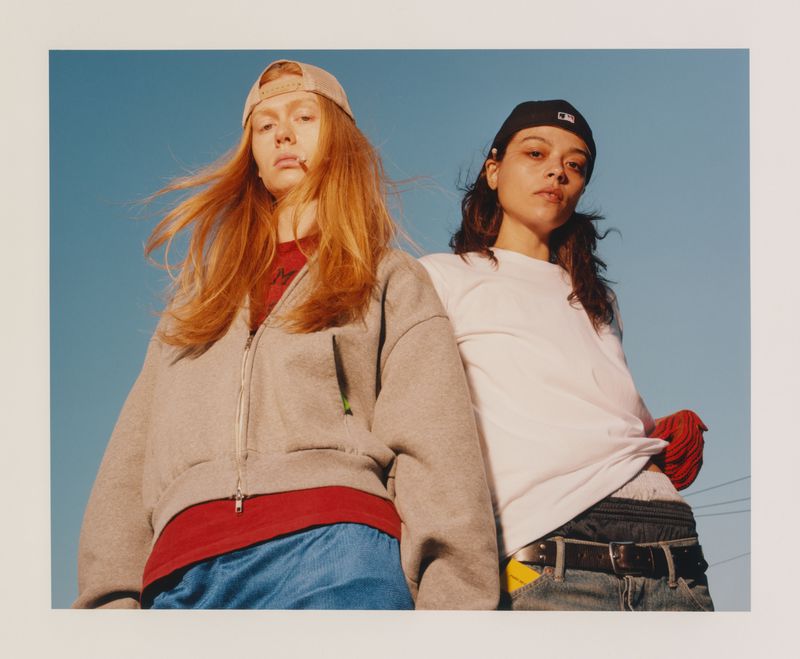 Low-angle shot of two women wearing casual streetwear and backward baseball caps against a clear blue sky.