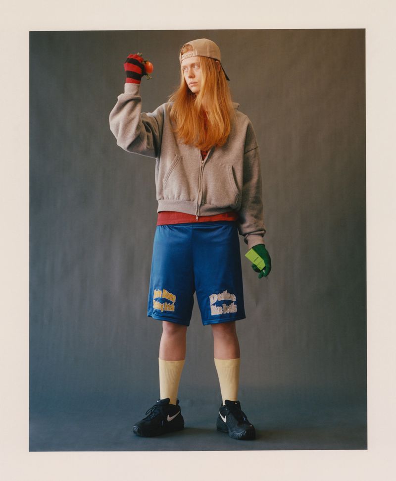 Model with long reddish hair wearing a grey zip hoodie, blue athletic shorts, cap, and mismatched gloves against a grey backdrop.