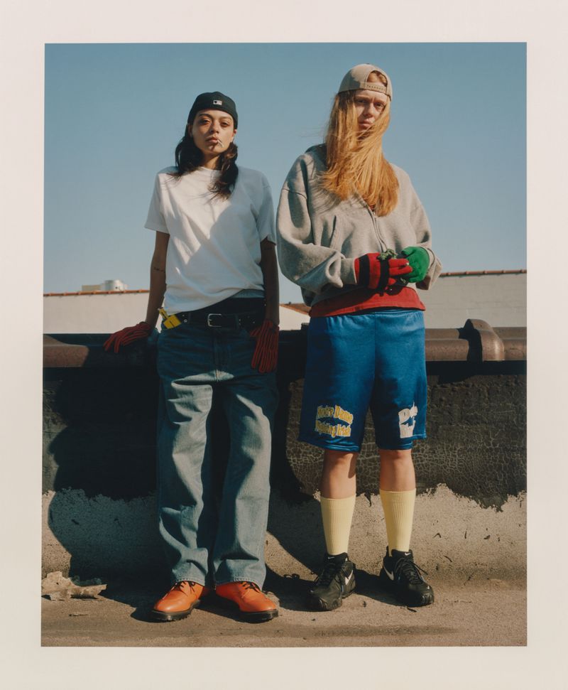Two models posing in streetwear with baggy jeans and athletic shorts on a sunny rooftop against a clear blue sky.