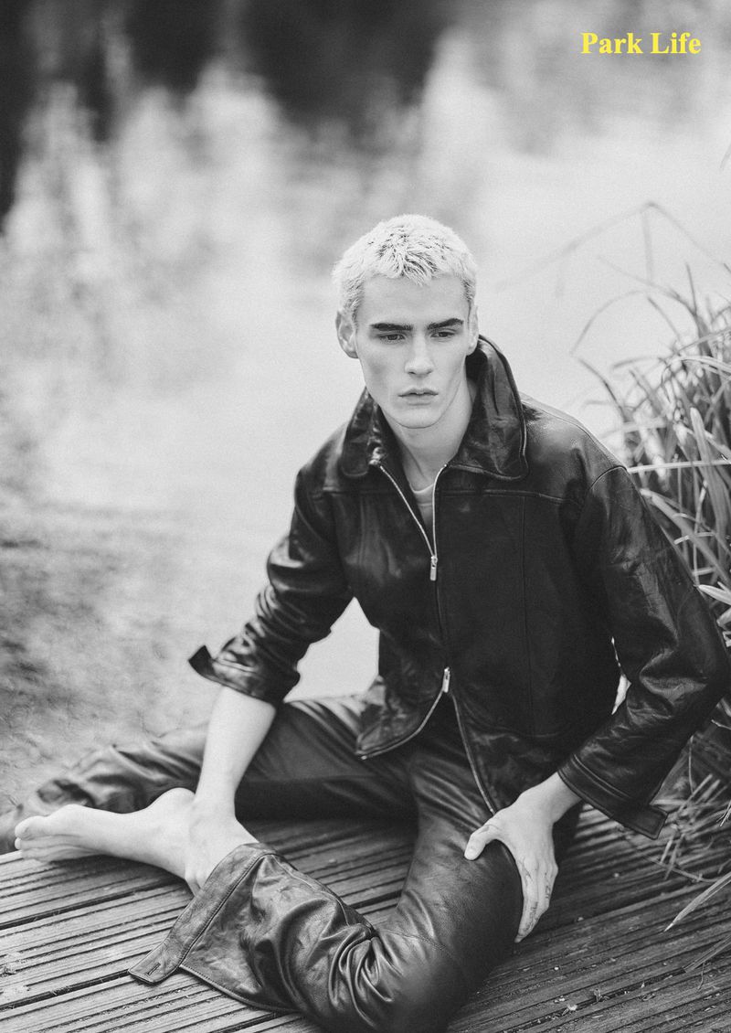 Bleach-blonde male model sitting barefoot on a wooden dock wearing a black leather jacket and trousers in a monochrome park setting.