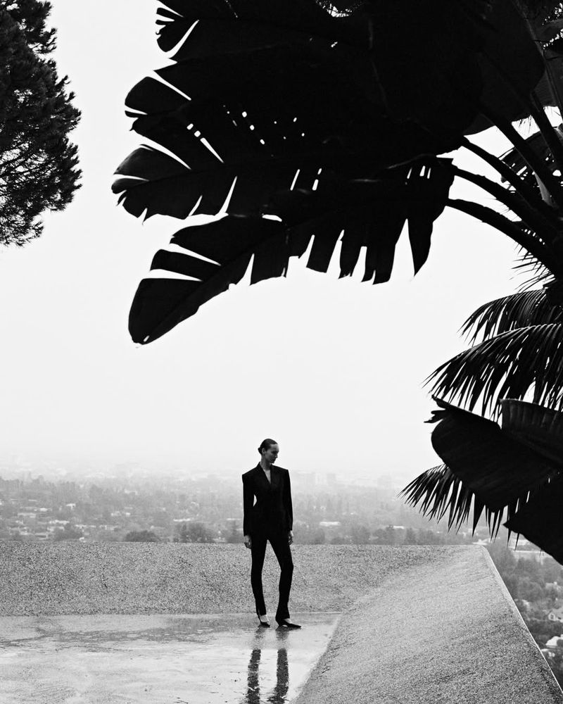 Julia Bergshoeff in a black tailored suit standing on a concrete rooftop under silhouetted palm fronds overlooking a valley.