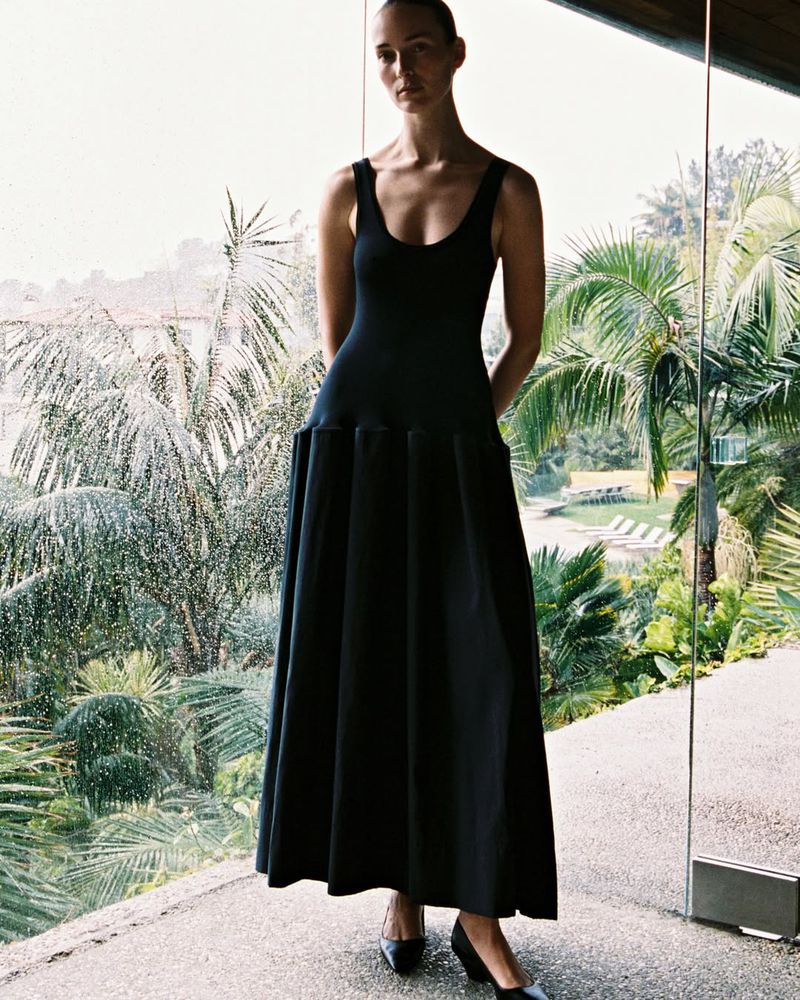 Model Julia Bergshoeff standing indoors wearing a sleeveless black drop-waist maxi dress in front of windows overlooking palm trees.