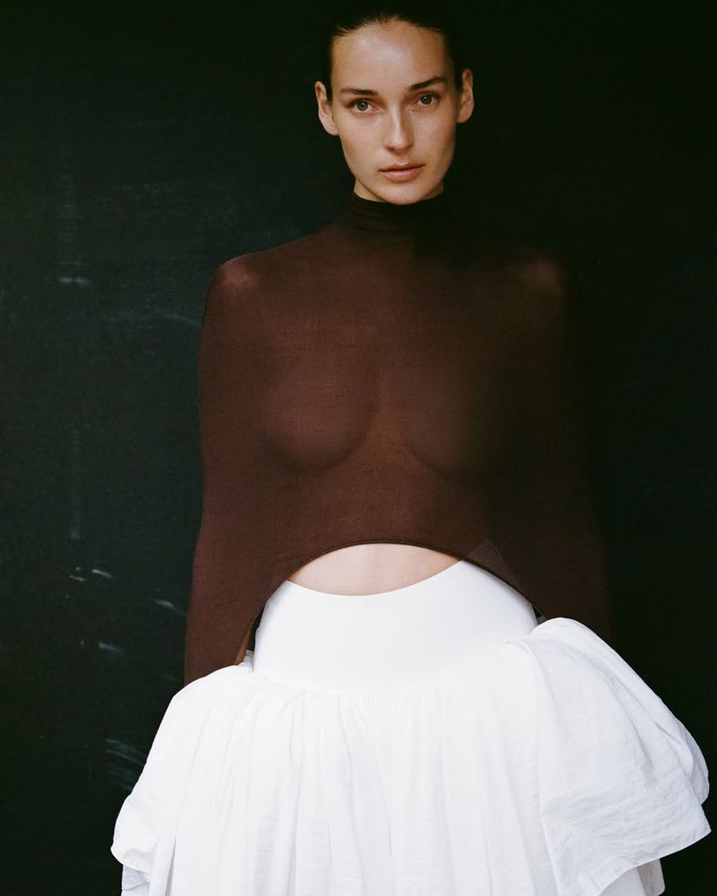 Julia Bergshoeff posing in a sheer brown high-neck top and a voluminous white tiered skirt against a dark black backdrop.