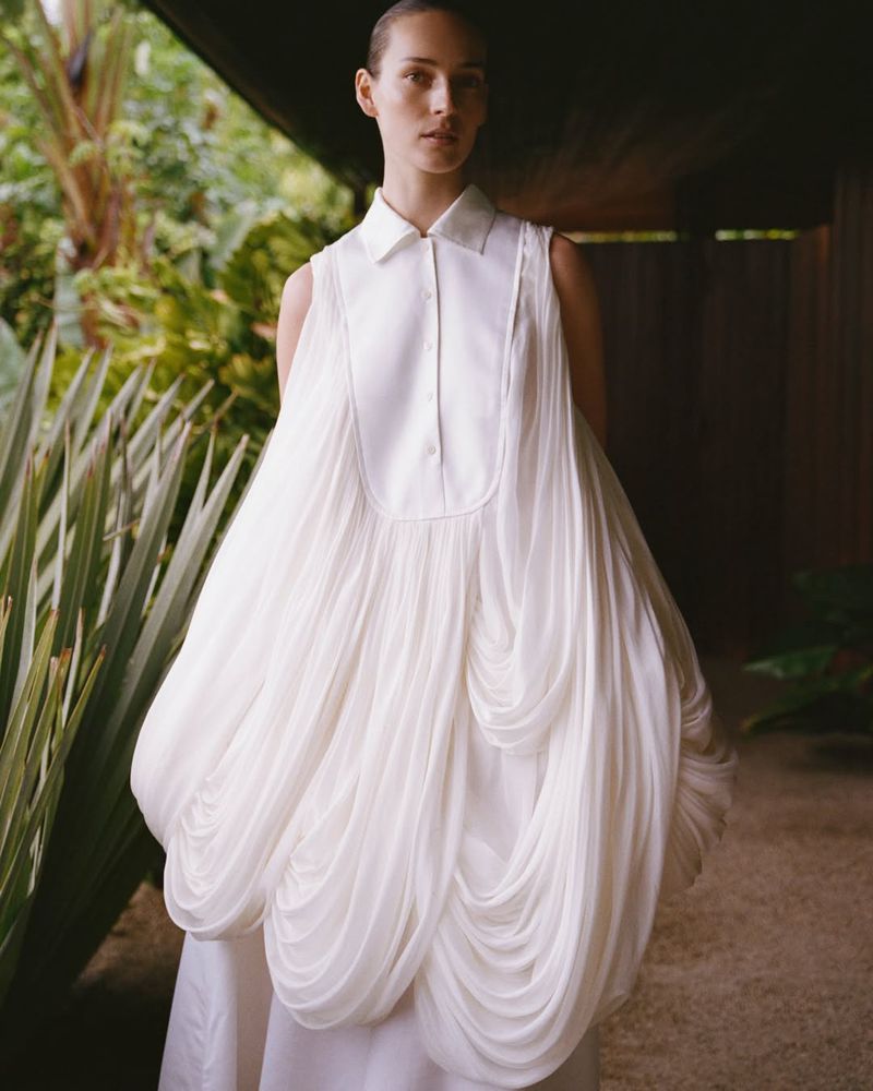 Model Julia Bergshoeff wearing a voluminous draped white sleeveless dress with a collared bodice in an outdoor garden setting.
