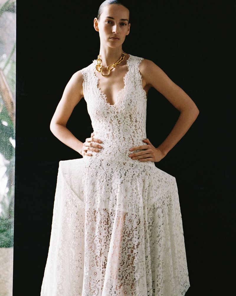 Model Julia Bergshoeff in a white lace dress with structured hips and a chunky gold necklace against a dark background.
