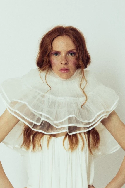 Close-up portrait of a red-haired woman wearing a large white ruffled collar and dress, against a white studio backdrop.