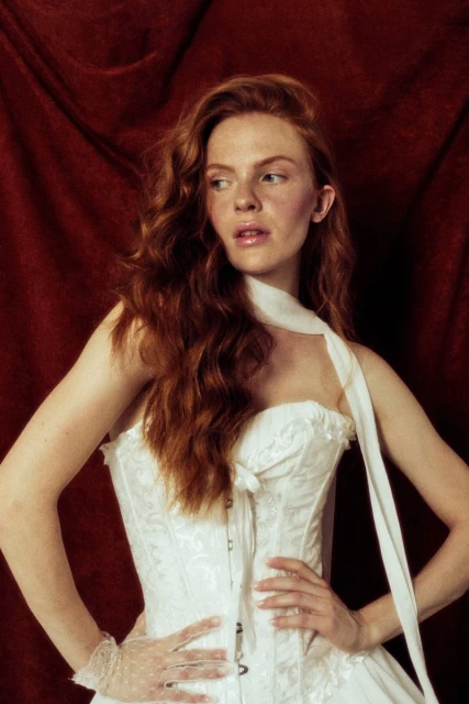 Red-haired woman in white corset dress poses against dark red backdrop in a fashion editorial image with soft, diffused lighting.