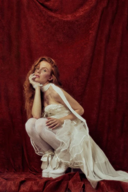 Red-haired woman in white dress crouching against a red velvet backdrop, wearing white platform shoes, with her hand on her chin.