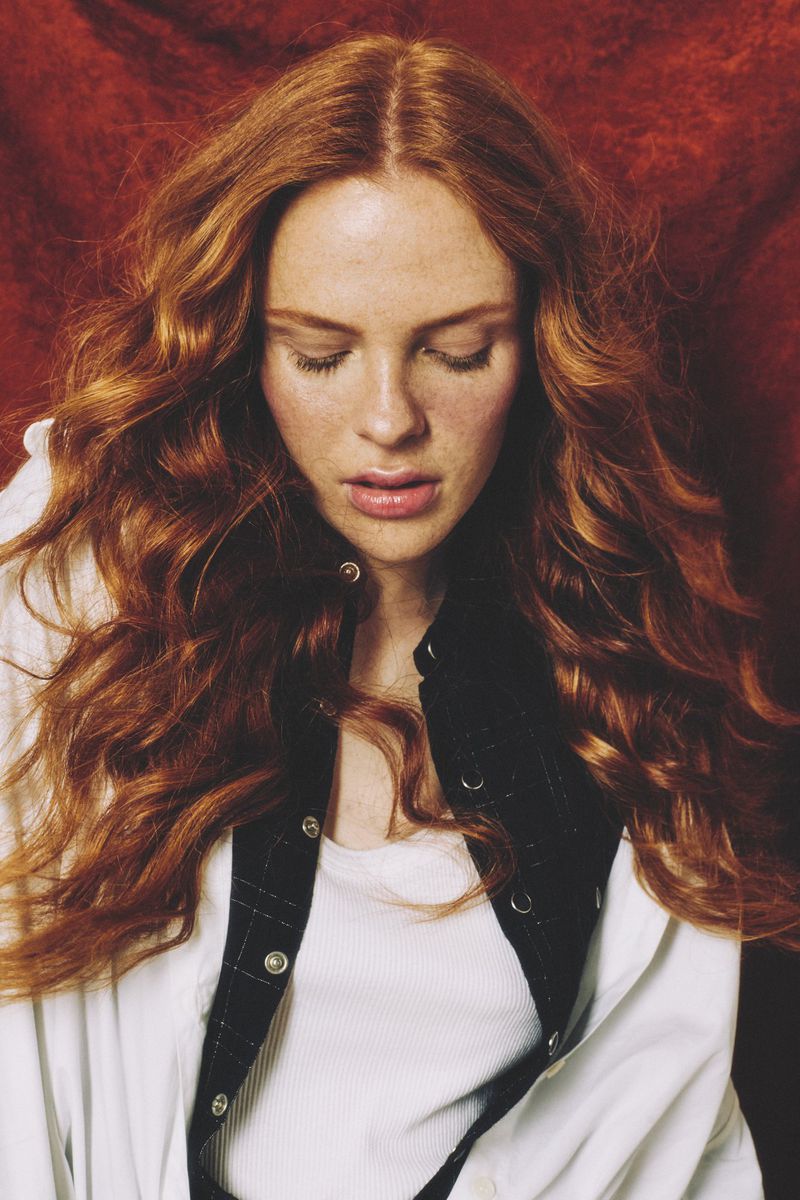 Close-up studio portrait of a young adult woman with long red wavy hair wearing a white buttoned shirt and black vest against a red backdrop.