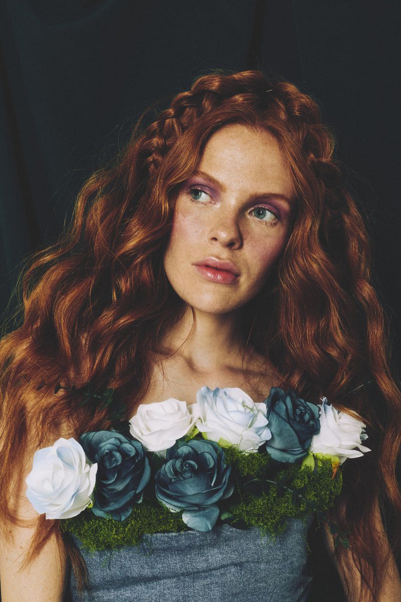 Portrait of a red-haired woman with braided accents, wearing a floral collar of blue and white roses against a dark backdrop.
