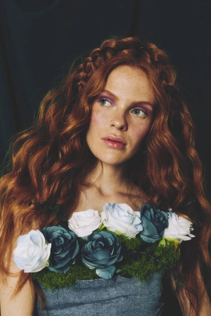 Portrait of a red-haired woman with braided accents, wearing a floral collar of blue and white roses against a dark backdrop.