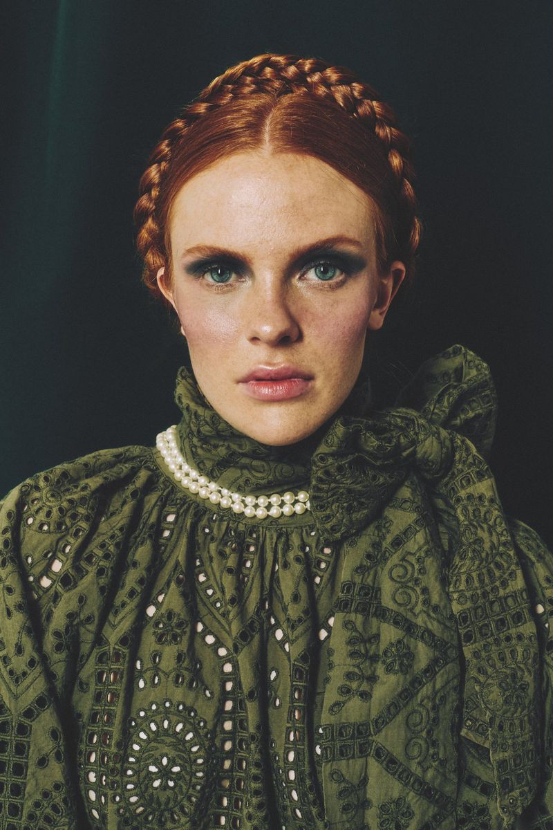 Close-up portrait of a red-haired woman in green dress with pearl necklace. The model has a braided hairstyle and dramatic eye makeup.