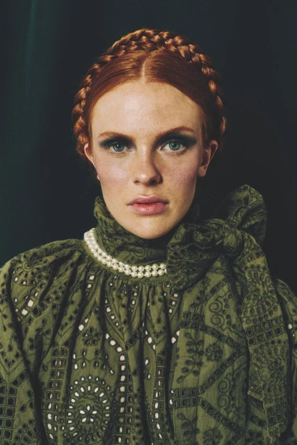 Close-up portrait of a red-haired woman in green dress with pearl necklace. The model has a braided hairstyle and dramatic eye makeup.