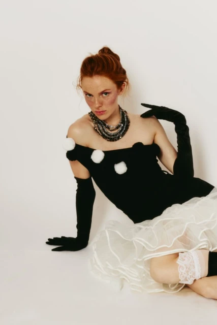 A red-haired woman sits on a white surface wearing a black off-the-shoulder dress with white pom poms, long gloves, jewelry, and lace-trimmed stockings.