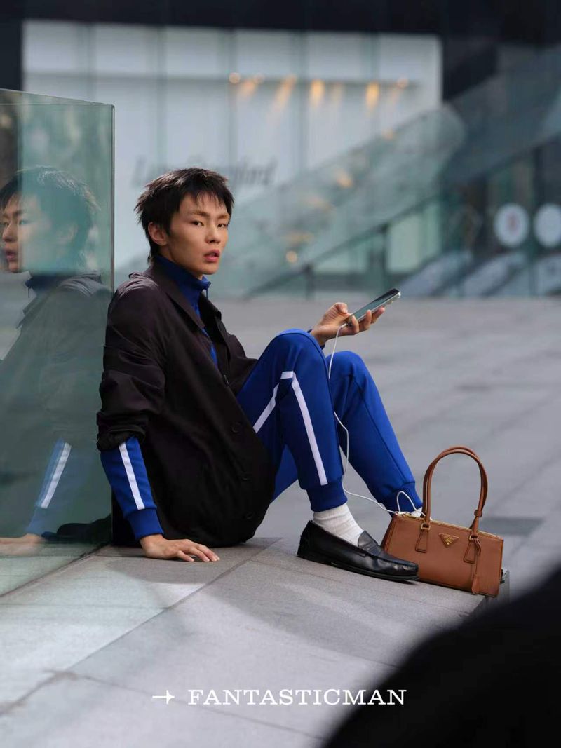 Model sitting on an urban ledge in a blue tracksuit and black overshirt holding a phone beside a structured brown leather handbag.