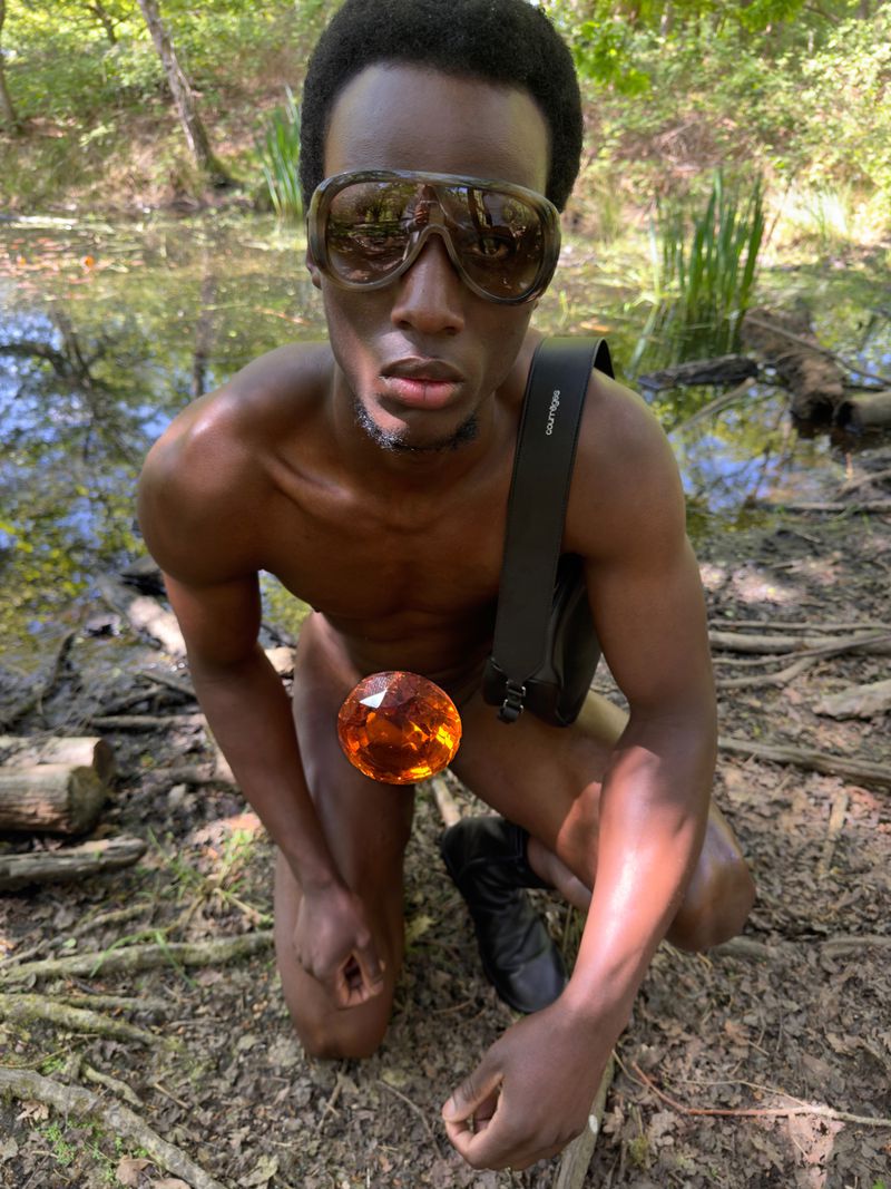 A Black man kneels outdoors in a woodland setting wearing large tinted wrap-around sunglasses and a black crossbody strap.