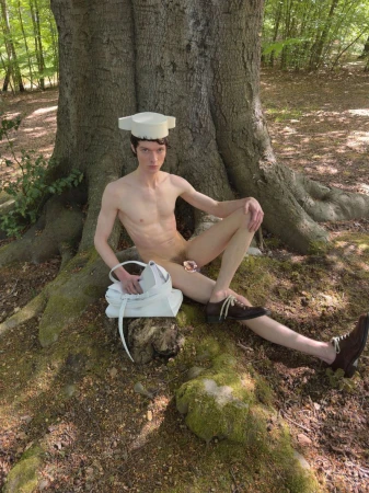 Model wearing a white structured hat and brown lace-up shoes seated against a large tree in a forest setting for Pull Letter Magazine
