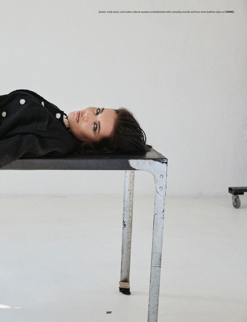 A woman lying flat on a dark industrial metal table wearing a black jacket against a plain white studio background.
