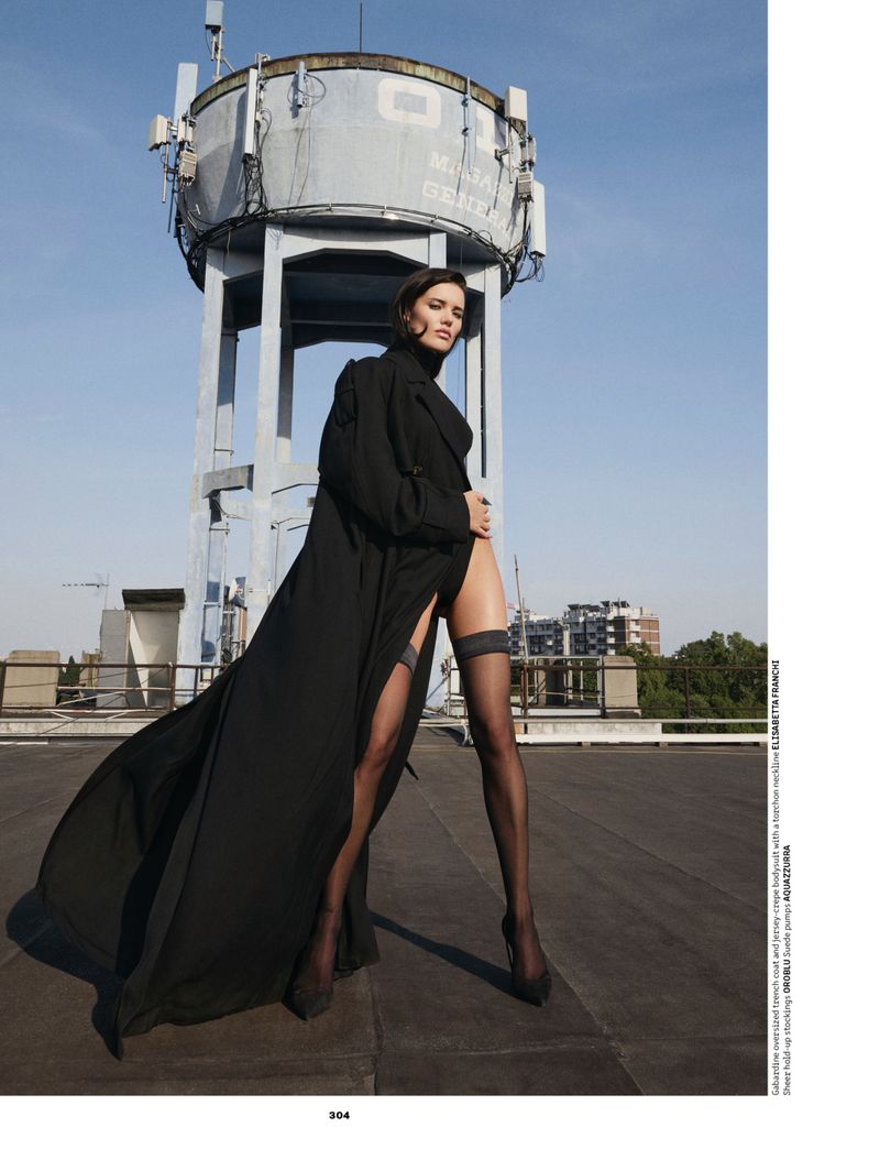 Model posing on a rooftop in a sweeping black coat, black bodysuit, and sheer thigh-high stockings before a tall water tower.