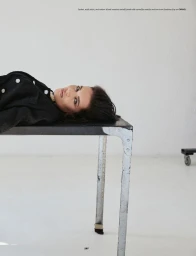 A woman lying flat on a dark industrial metal table wearing a black jacket against a plain white studio background.