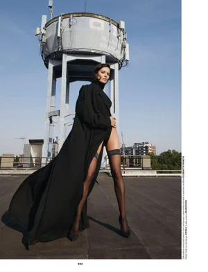 Model posing on a rooftop in a sweeping black coat, black bodysuit, and sheer thigh-high stockings before a tall water tower.