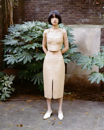 Model Asako Sato in a beige quilted crop top and matching split midi skirt posing outdoors by foliage and brick wall.