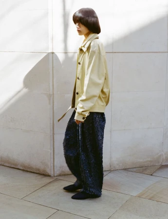 Model Asako Sato in a beige jacket and textured dark trousers standing in profile against a sunlit stone wall.