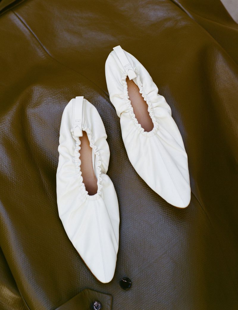 Pair of off-white pointed-toe gathered leather ballet flats resting on an olive brown textured leather surface.