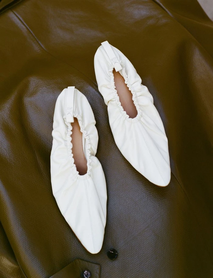 Pair of off-white pointed-toe gathered leather ballet flats resting on an olive brown textured leather surface.