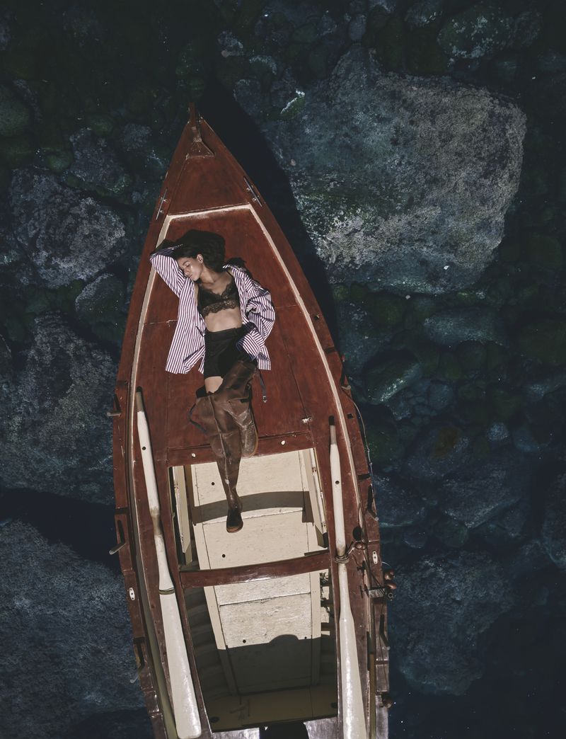 High-angle view of model Jennifer Matias lying inside a wooden rowing boat on clear water over submerged rocks.
