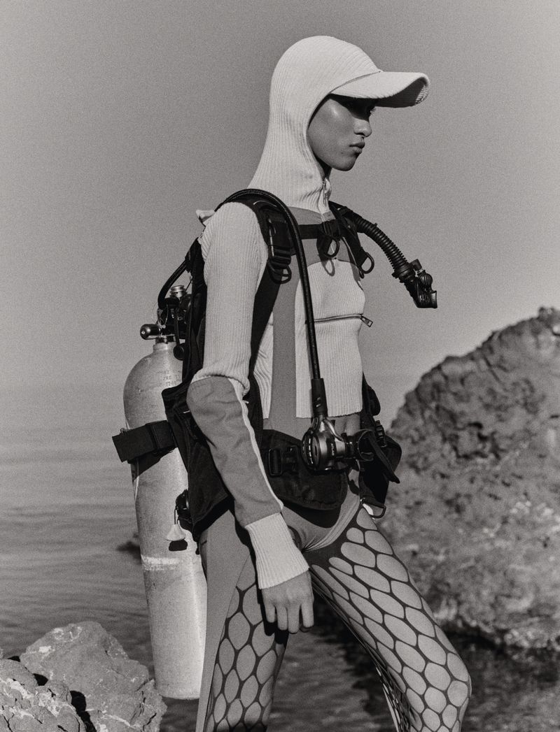 Model wearing a scuba tank, diving harness, and knit visor hood standing on a rocky shoreline in black and white.