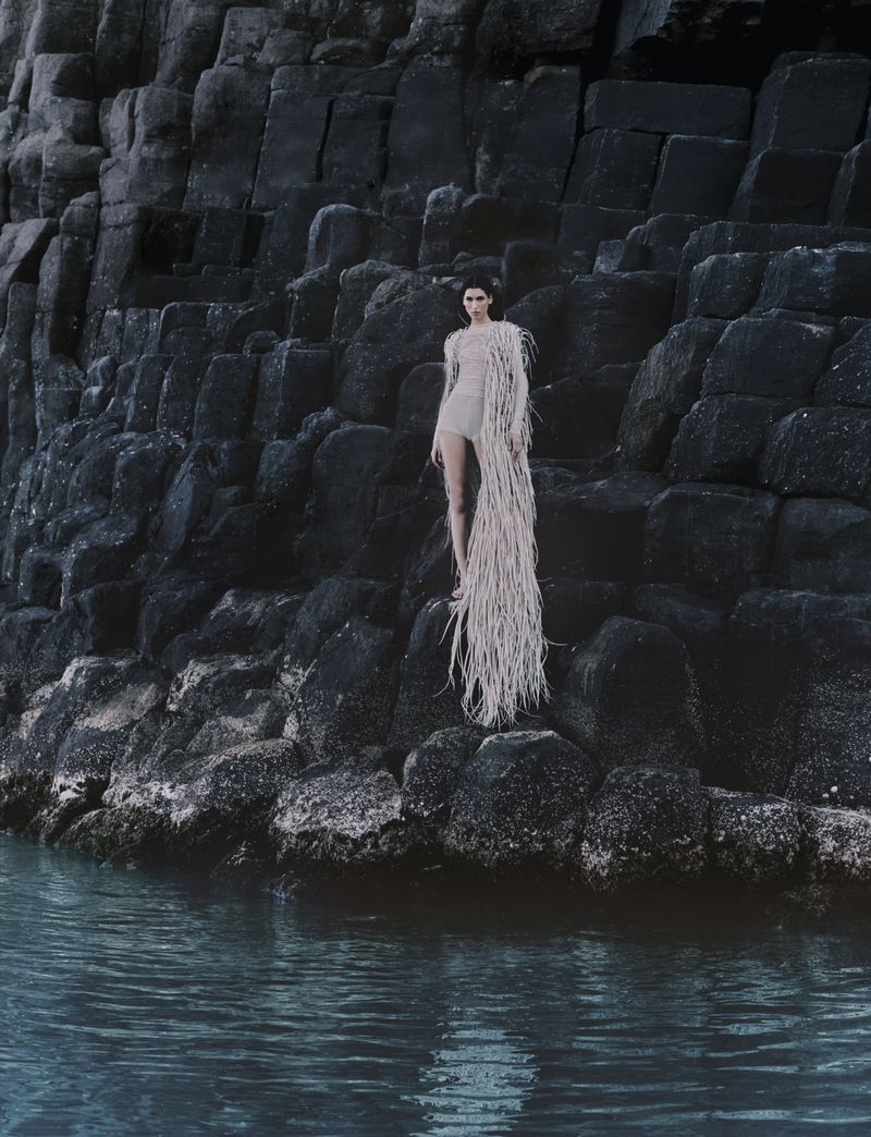 Model Jennifer Matias in a long textured fringed bodysuit standing on dark coastal basalt rocks above the sea water.