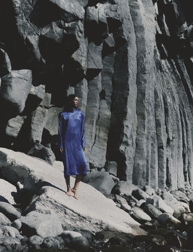 Model Jennifer Matias wearing a long blue dress while standing barefoot against towering basalt rock columns on a rocky coast.