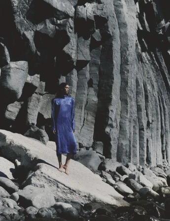 Model Jennifer Matias wearing a long blue dress while standing barefoot against towering basalt rock columns on a rocky coast.