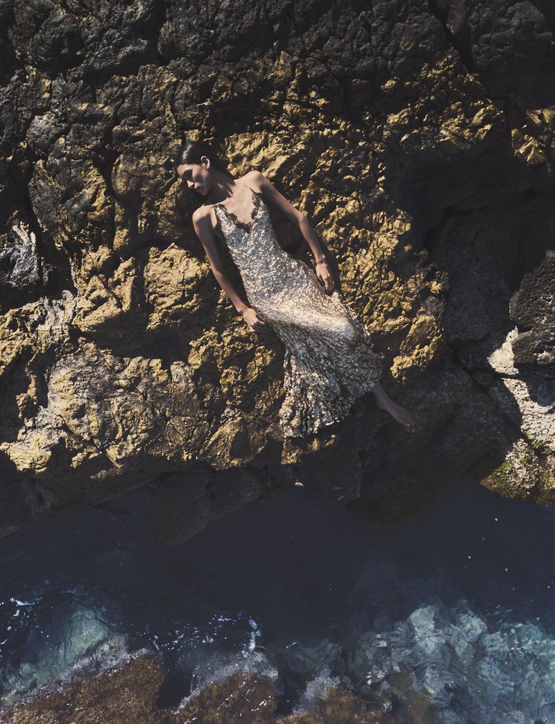 Model Jennifer Matias wearing a sparkling silver sequined gown while reclining on a rocky cliff edge above ocean water.