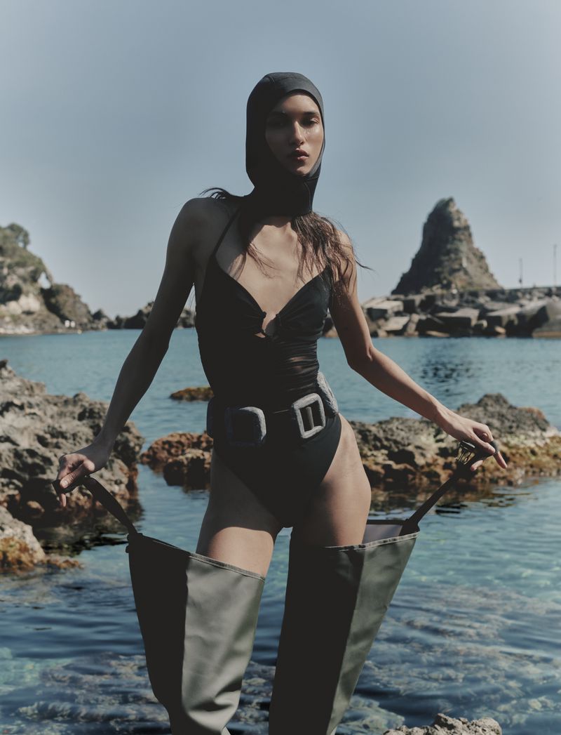 Model Jennifer Matias stands on coastal rocks wearing a black swimsuit, fitted headpiece, and thigh-high wader boots.