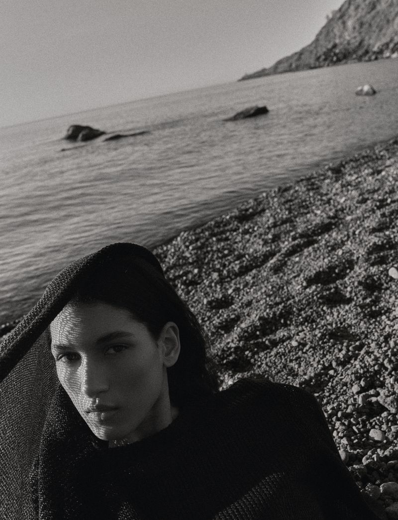 Monochrome close-up of model Jennifer Matias draped in a black textured knit shawl on a rocky pebble beach overlooking the sea.