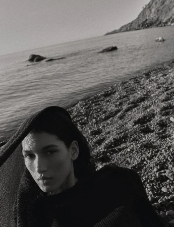 Monochrome close-up of model Jennifer Matias draped in a black textured knit shawl on a rocky pebble beach overlooking the sea.